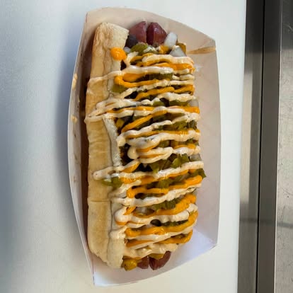 Weiner with relish, mustard and a white sauce drizzle in a paper tray
