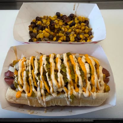 Loaded weiner with sauces and relish next to a tray of corn and black bean salad
